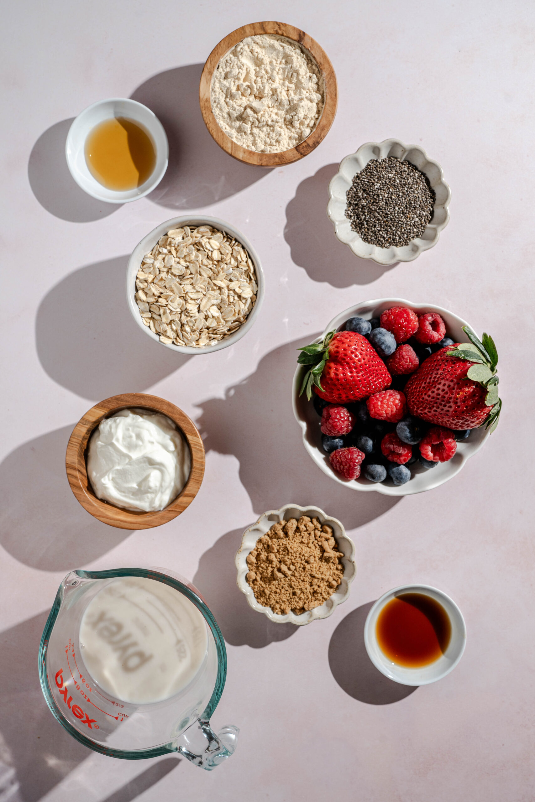 ingredients to make berry cheesecake overnight oats including almond milk, rolled oats, dairy free yogurt, protein powder, berries, chia seeds, vanilla extract, maple syrup, and gluten free graham crackers
